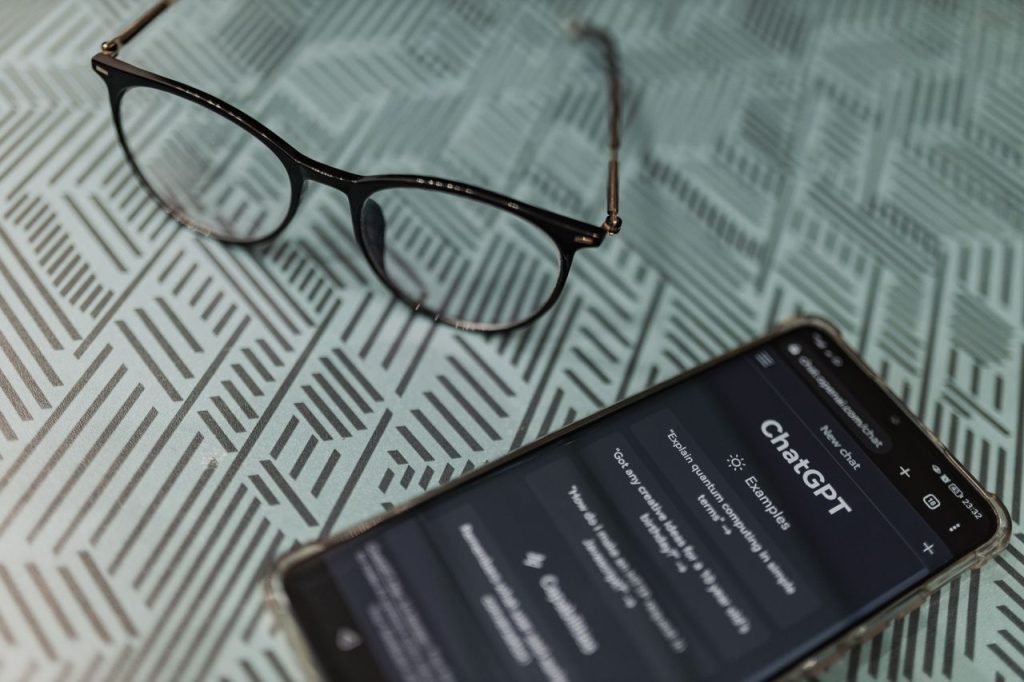 pexels photo 16094061 Eyeglasses next to a smartphone displaying the ChatGPT AI app on a patterned surface.